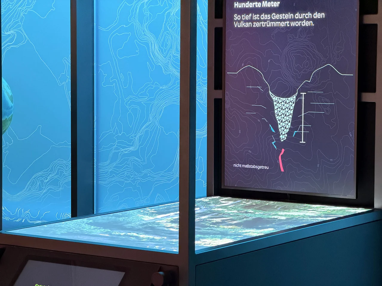 An exhibition installation with illuminated topographic walls, a vertical volcanic diagram and a projected tabletop.